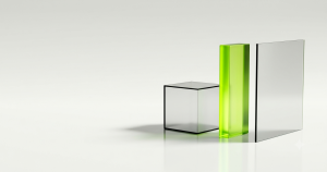 Three translucent glass objects of different shapes standing in a row on a clean reflective white floor: a solid cube, a tall narrow slab, and a thin wide sheet, two of them clear and softly frosted, one glowing with lime green